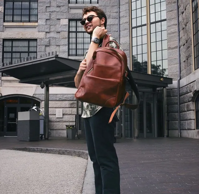 Man wearing sunglasses in front of stone building casually slinging brown leather backpack over shoulder