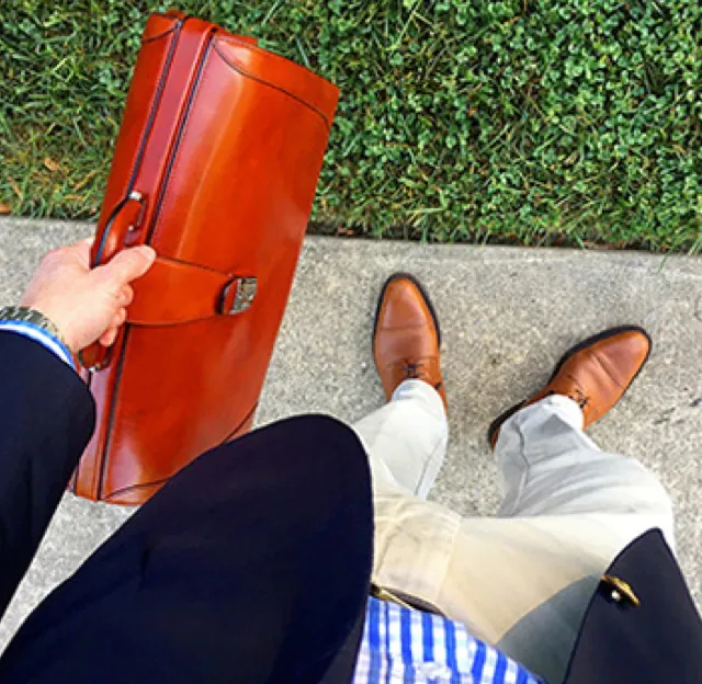 Perspective shot as if looking down along a man's body while he holds a light brown leather briefcase which match his brown leather shoes